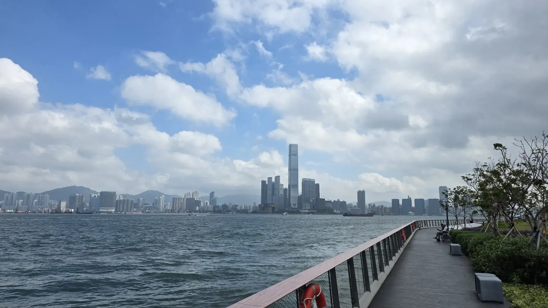 A Victoria Harbour promenade facing the Kowloon skyline, with the International Commerce Centre at the centre